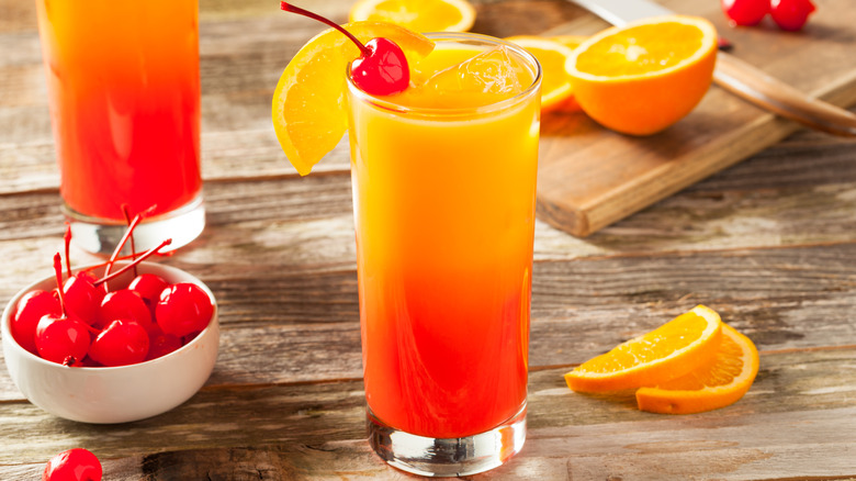 Orange and yellow Tequila Sunrise on a wooden board with a bowl of cherries and sliced orange.