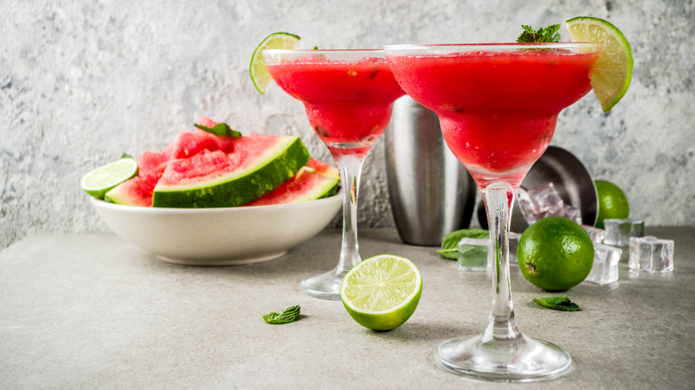 Two Margarita glasses filled with a vibrant pink cocktail and garnished with a lime next to a bowl of sliced watermelon on a concrete bar.