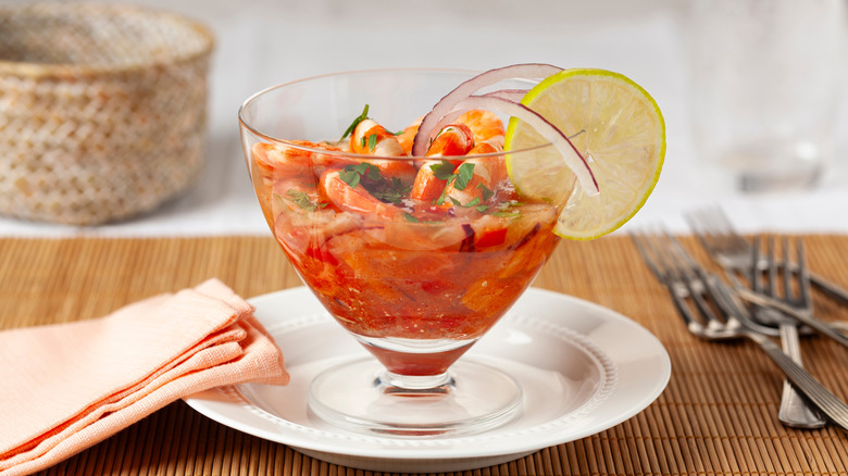 Cocktail dish of ceviche, red broth with large prawns,  on straw placement with cutlery