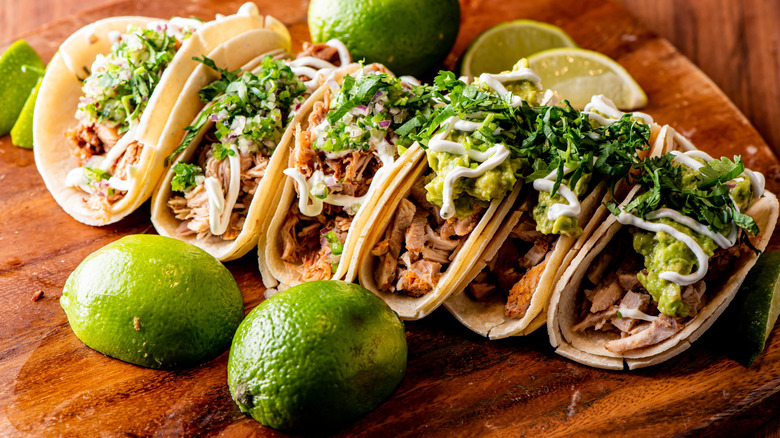 Row of meat tacos on wooden cutting board surrounded by limes