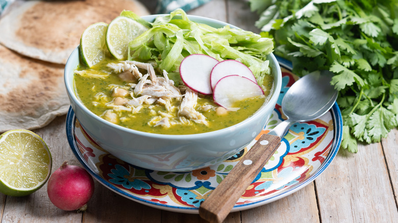 White bowl of pozole on Talavera plate, with wood-handled spoon, and bunch of cilantro