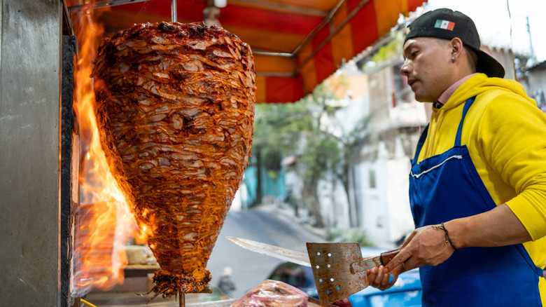 Man at roadside stand carving carne al pastor from trompo