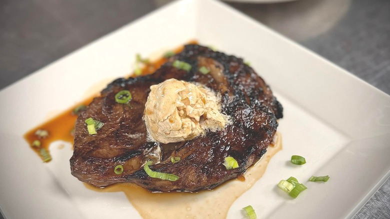 A butter and scallion-topped steak on a square, white plate.