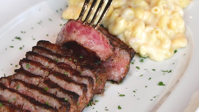 A fork lifting a slice of steak from a white plate holding more slices and macaroni and cheese.