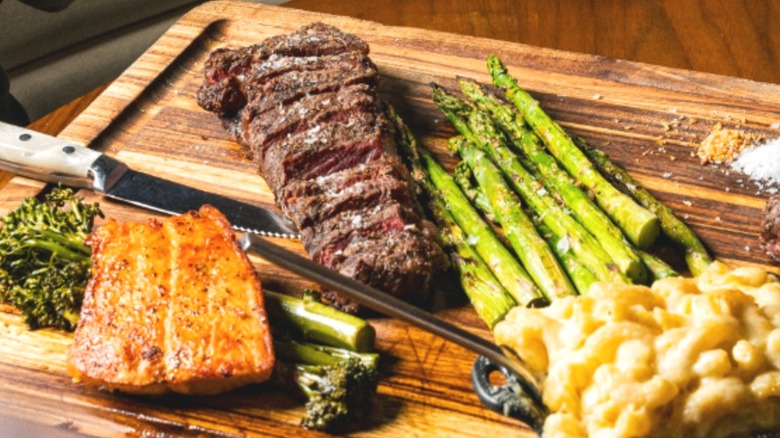 Sliced steak, salmon, asparagus, and macaroni and cheese on a wooden cutting board.