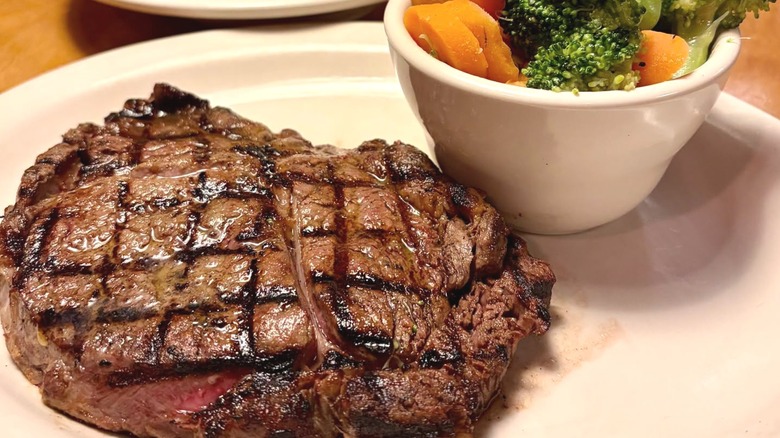 A large steak and a cup of vegetables positioned on a white plate.