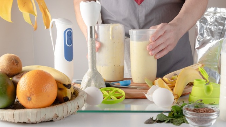 Person stands by a blender of orange shake liquid with basket of fruits to the side