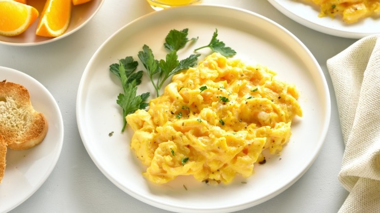 White plate of scrambled eggs with green garnish surrounded by plates of fruit and bread