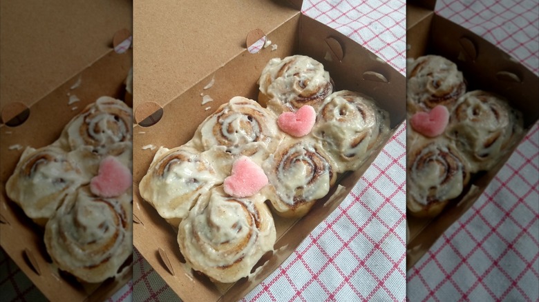 Six cinnamon rolls covered in cream cheese with two pink heart candies