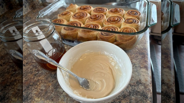 Tray of cinnamon rolls next to glass jar of maple syrup and bowl of frosting