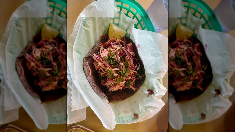 Green plastic basket, lined with white wrapper, holds a taco with a dark blue tortilla filled with slaw.