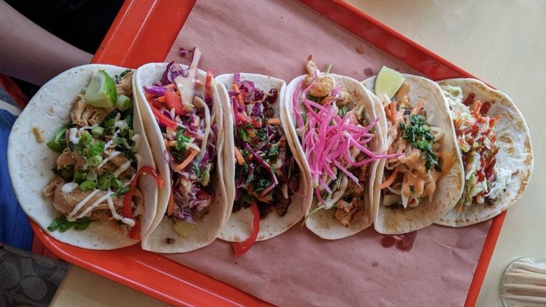 Six flour tortilla tacos with various colorful fillings, served on a red plastic tray