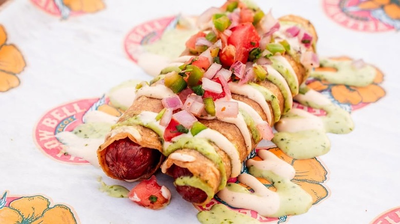 Two hot dogs wrapped in a fried corn tortilla and drizzled with green and white sauces and pico de gallo.