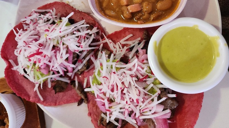 Pink tortilla tacos filled with meat, avocadoes and shredded radishes, on a plate with a bowl of beans and a bowl of creamy green salsa