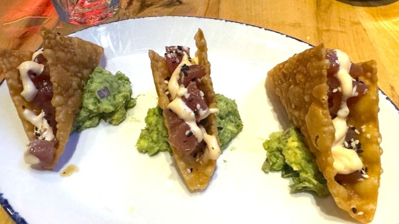 Three tiny tacos made of fried wanton wrappers, filled with poke, sitting on a pile of guacamole.