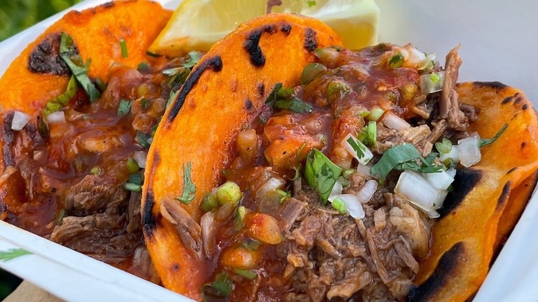 Two birria tacos in a takeout container with lemon