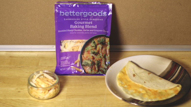 A bag of Bettergoods Gourmet Baking Blend beside a dish of the unbagged cheese and a small quesadilla on a plate
