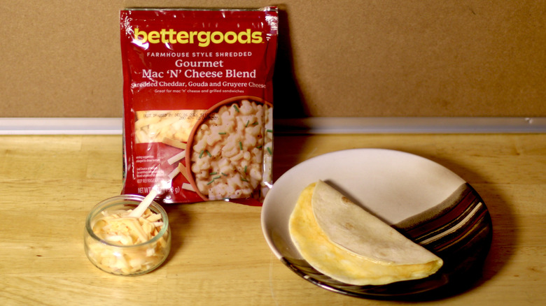 A bag of Bettergoods Gourmet Mac 'N' Cheese Blend beside a dish of the unbagged cheese and a small quesadilla on a plate