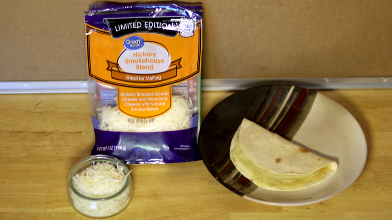 A bag of Great Value Limited Edition Hickory Smokehouse Blend shredded cheese beside a dish of the unbagged cheese and a small quesadilla on a plate