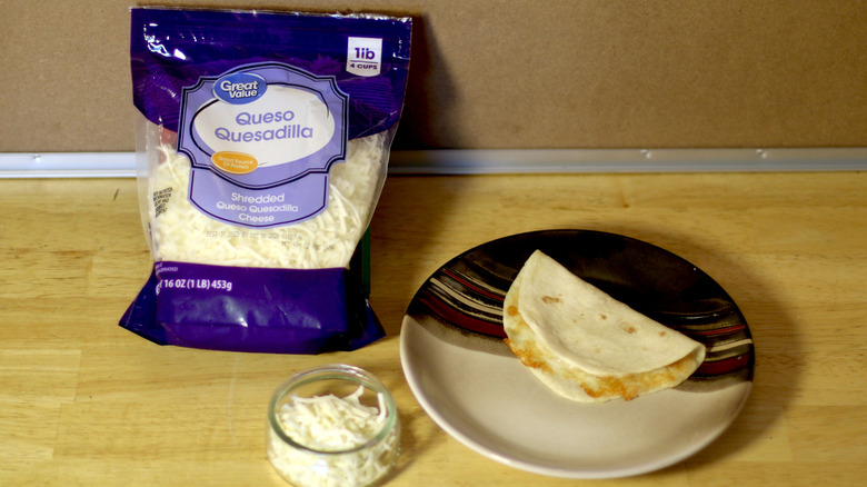 A bag of Great Value Queso Quesadilla shredded cheese beside a dish of the unbagged cheese and a small quesadilla on a plate