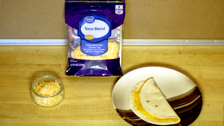 A bag of Great Value Taco Blend shredded cheese beside a dish of the unbagged cheese and a small quesadilla on a plate