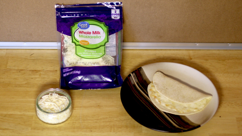 A bag of Great Value Whole Milk Mozzarella shredded cheese beside a dish of the unbagged cheese and a small quesadilla on a plate