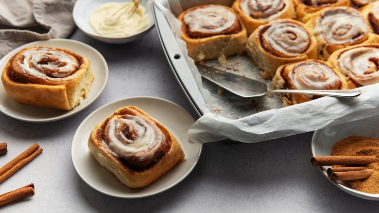 a pan of cinnamon rolls next to individual rolls on plates with cinnamon sticks and icing in the background