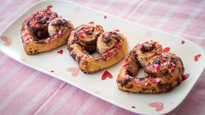 three heart shaped cinnamon rolls with pink and red sprinkles on a white plate with pink and red hearts on a pink and white striped tablecloth