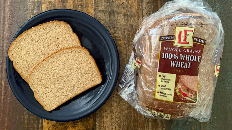 Two slices of L'oven Fresh 100% Whole Wheat Wide Pan bread next to a loaf of the same bread