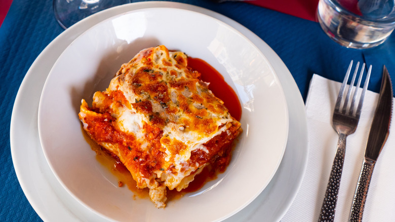 Piece of lasagna with watery sauce in a white bowl on a white plate with a fork and knife on a white paper napkin on the side