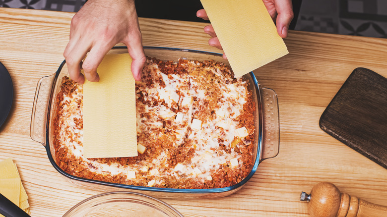 Hands putting lasagna sheets in casserole dish atop meat sauce and cheese