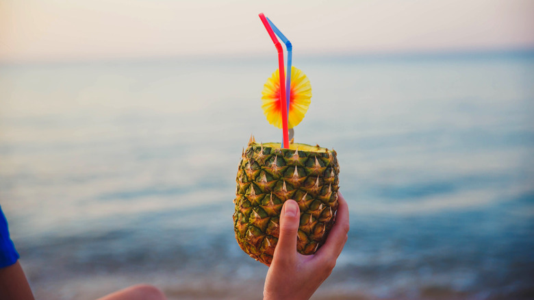 A woman's hand holding a pineapple with two straws and a drink umbrella up against the ocean stretching to the horizon.