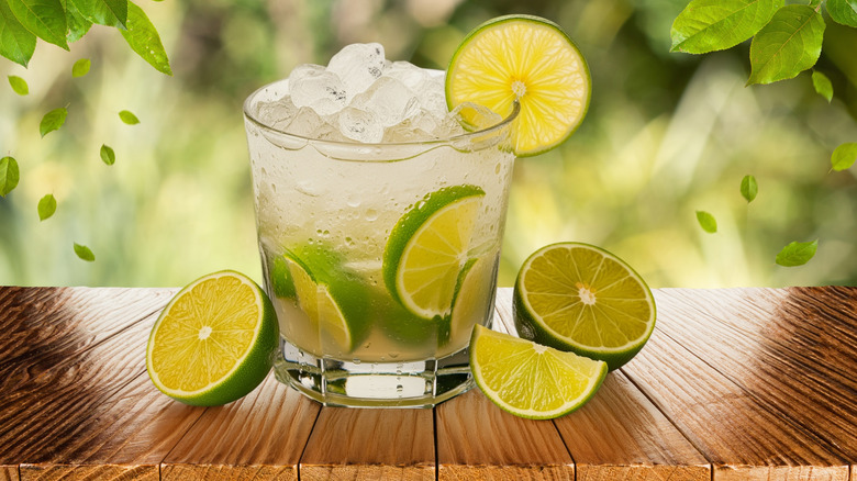 A rocks glass filled with a clear beverage and garnished with several limes on a wooden table.