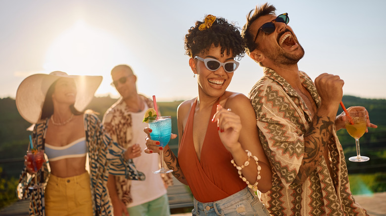 A group of four friends laughing and enjoying colorful drinks outside on a sunny day.