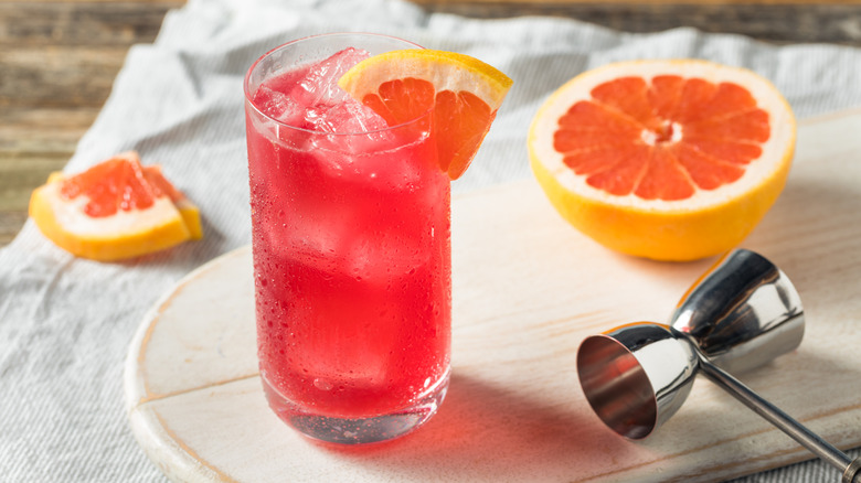 A pink liquid in an ice-filled glass next to half a grapefruit on a cutting board.