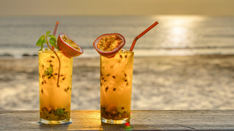 Two highball glasses filled with a light orange liquid with the sun setting over the ocean in the background.