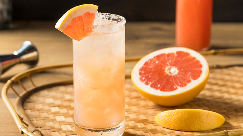 A highball glass with a light orange liquid, garnished with a wedge of grapefruit, on a bamboo tray with half a grapefruit.