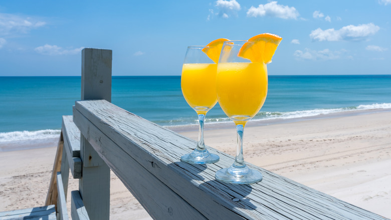 Two wine glasses filled with bright orange liquid, on a rail with an empty beach and deep blue ocean in the background.