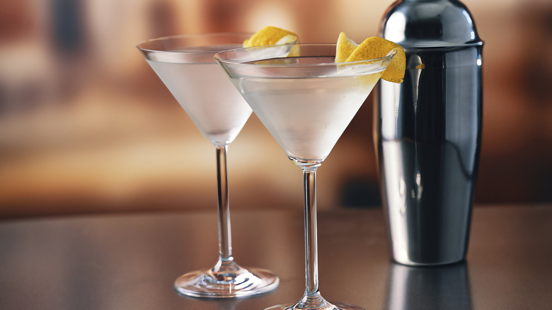 Two frosted martini glasses with a twist of lemon next to a metallic cocktail shaker.