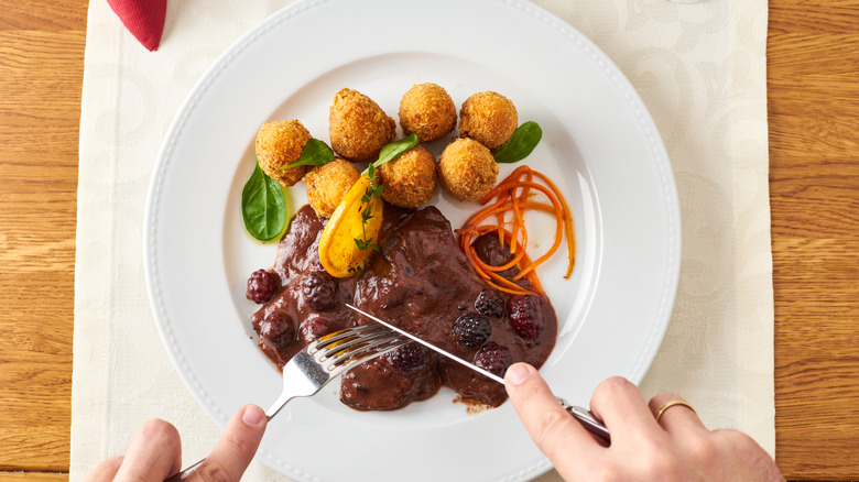 Person cuts into a cut of meat smothered in mulberry sauce on plate with croquettes