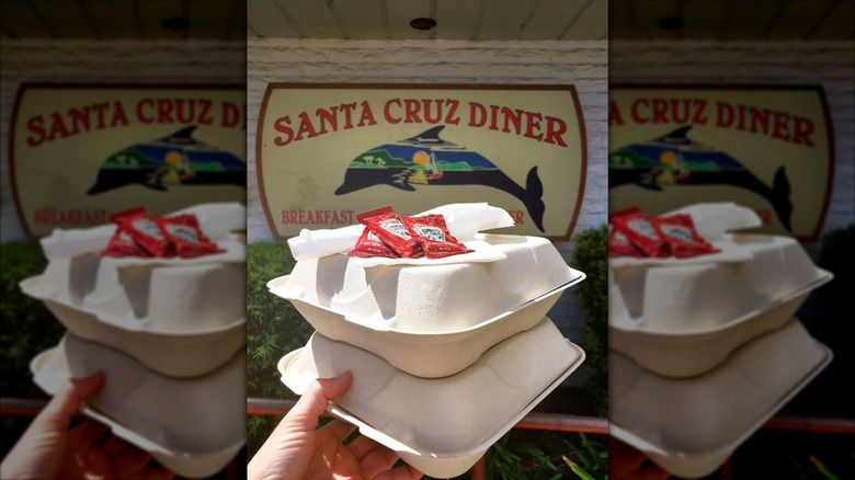 Hand holding two takeout boxes topped with ketchup sachets in front of Santa Cruz Diner sign