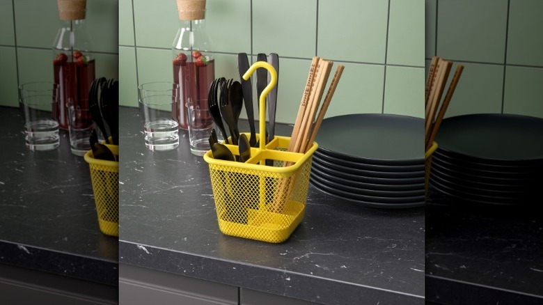 yellow Uppdatera flatware caddy holding forks, spoons, and chopsticks sitting on a black kitchen counter next to glasses, a liquid-filled bottle, and a stack of black plates