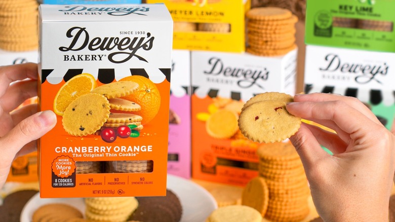 Dewey's Bakery cookies assorted boxes in background and cranberry orange box in front with hand holding a cookie