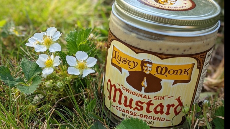 Lusty Monk Mustard Original Sin mustard jar on grass with white flower beside it
