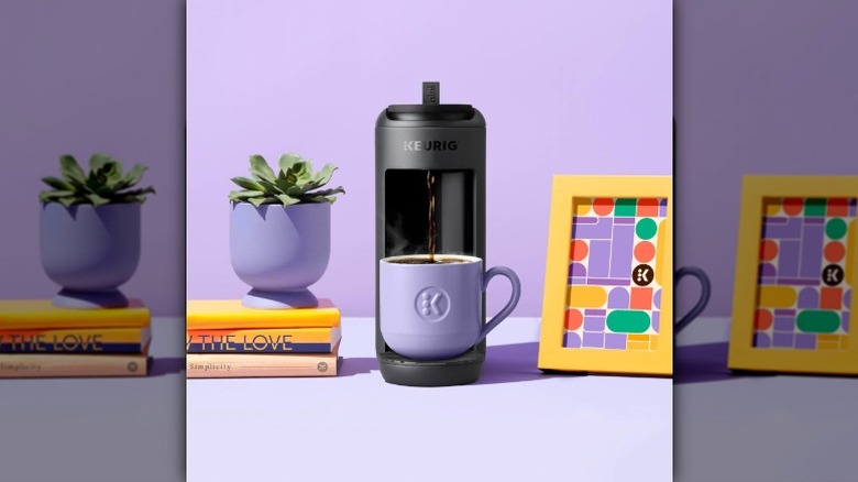 Black K-Mini Mate coffee maker with a purple coffee cup, books, plant in purple pot, and framed print