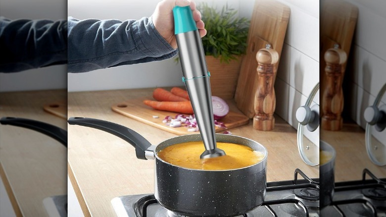 hand holding a cyan blue Turelar immersion blender in a pot of orange pureed soup