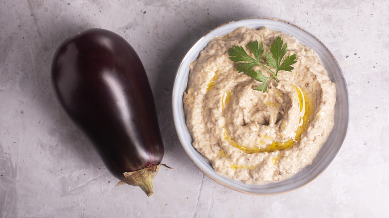 a bowl of Baba Ghanoush drizzled with olive oil and topped with flat-leaf parsley next to an eggplant