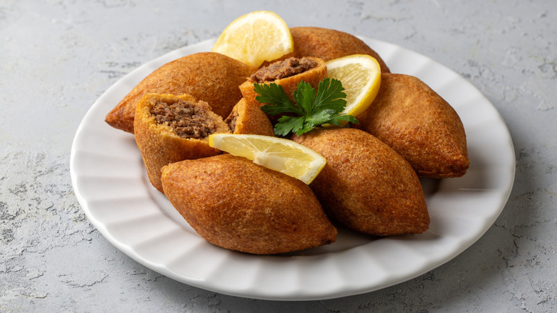 fried kibbeh on a plate with lemon wedges and flat-leaf parsley, one kibbeh split open to reveal ground meat