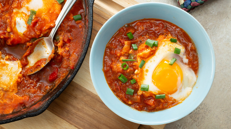 a bowl of Shakshuka with poached egg and chives served from a larger cast iron pan of shakshuka