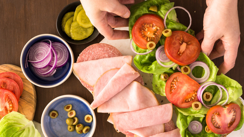 Hands assembling a sub with deli meat and fresh veggies